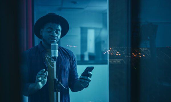 Portrait of Successful Young Black Artist, Singer, Performer Sin Portrait of Successful Young Black Artist, Singer, Performer Singing His Hit Song for the New Album. Wearing Stylish Hat, Holding Smartphone and Standing in Music Record Studio Soundproof Room.