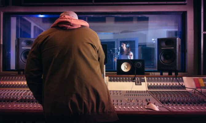 Sound engineer working at audio control panel with singer singing in recording room in background. Musicians producing music in professional recording studio.
